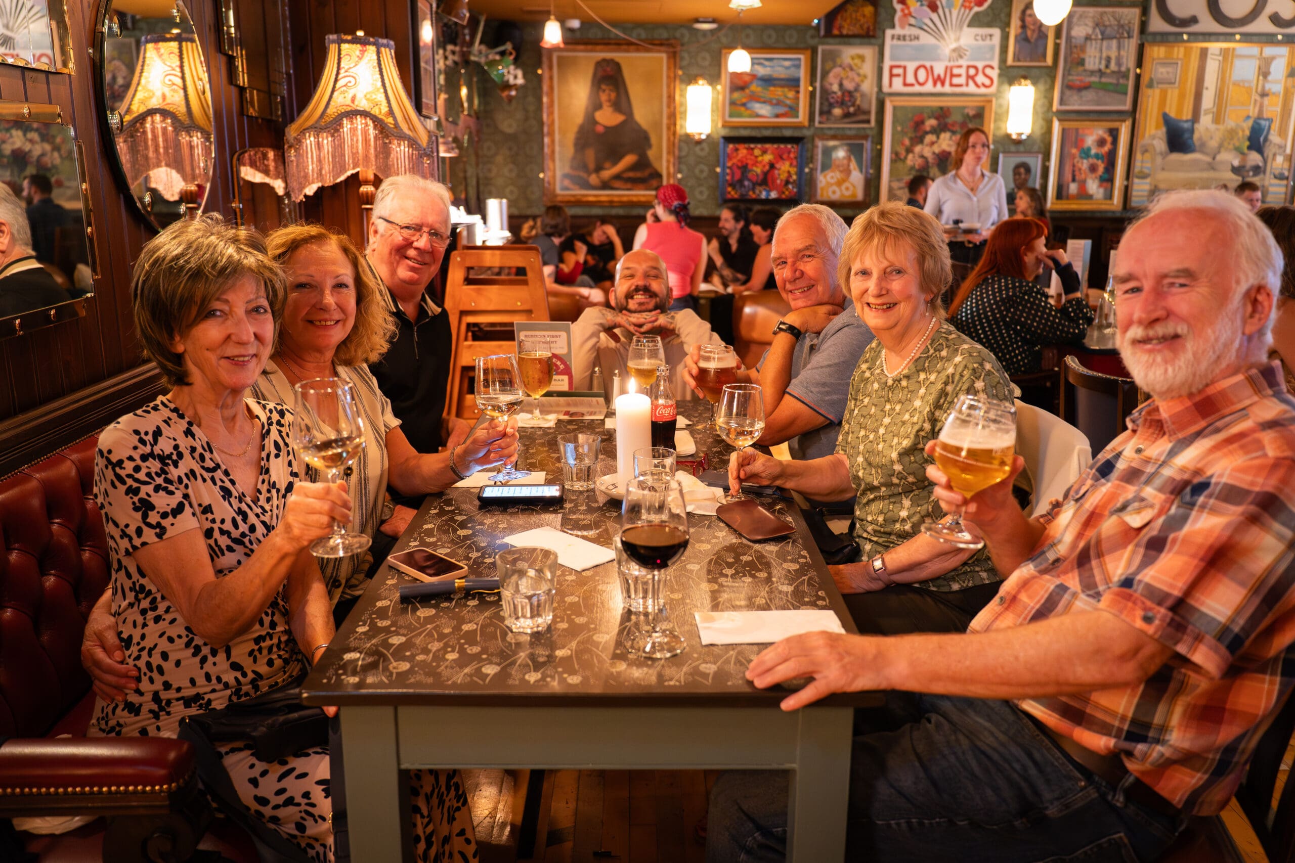 A group of seven smiling older adults sit around a table in a cozy pub, holding drinks and posing for a photo. The background features vintage lamps, paintings, colorful wall art, and a cheerful marinero theme.