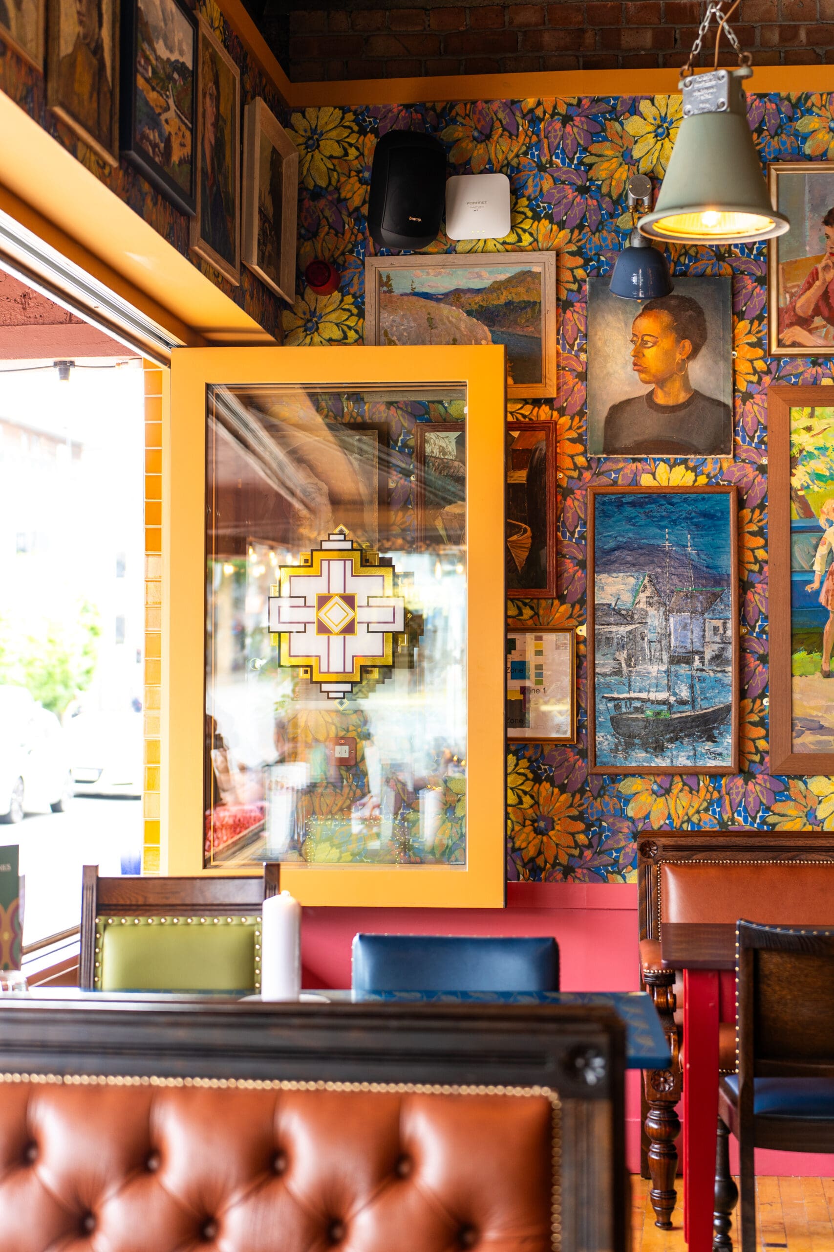 A brightly colored restaurant interior features leather seating, vibrant floral wallpaper, framed art, and a large yellow-framed window with decorative glass, creating a lively marinero atmosphere filled with natural light.