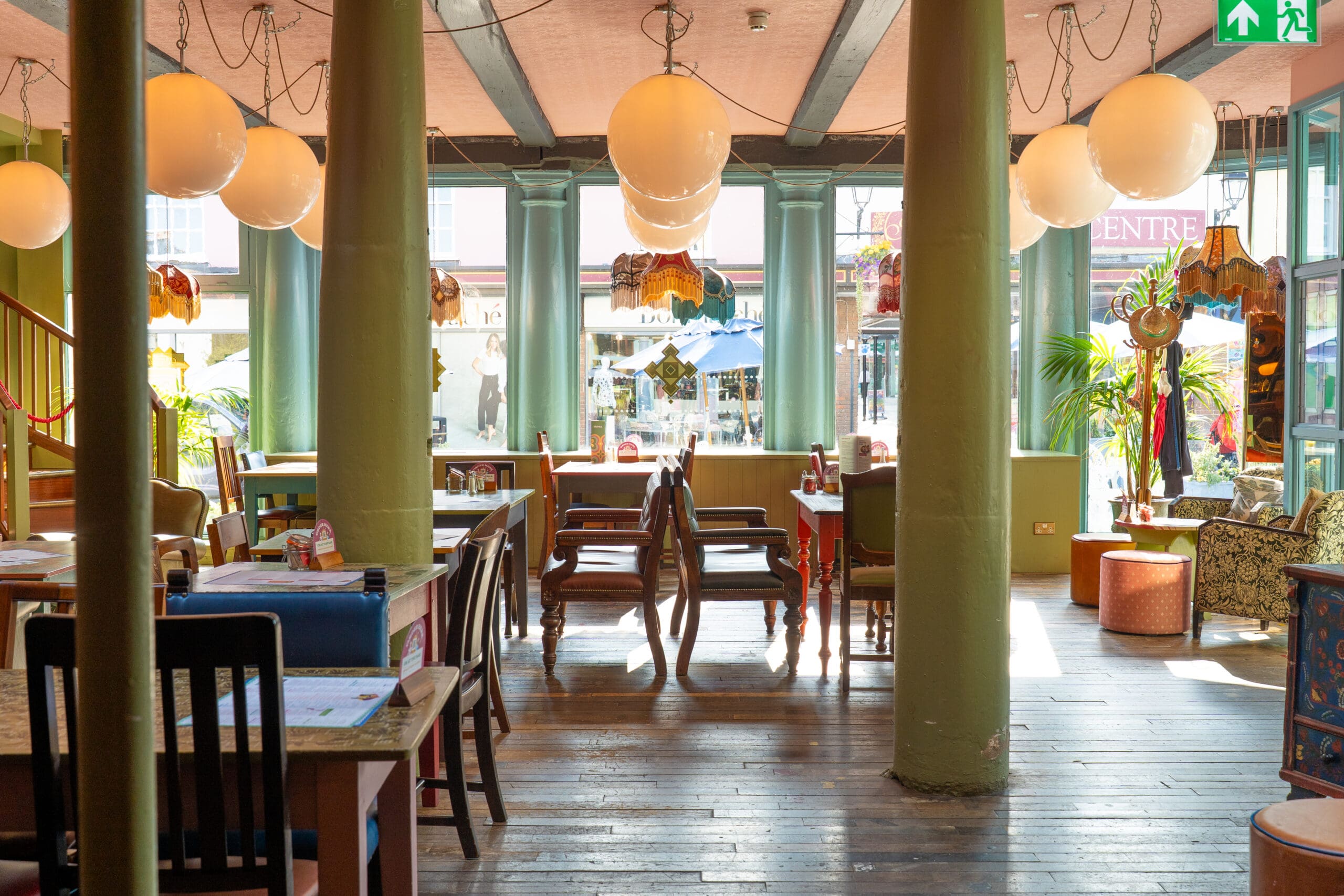 A bright, cozy café with mismatched chairs, wooden tables, and Bianco globe pendant lights. Large windows let in sunlight, illuminating green columns and eclectic decor inside the inviting space.