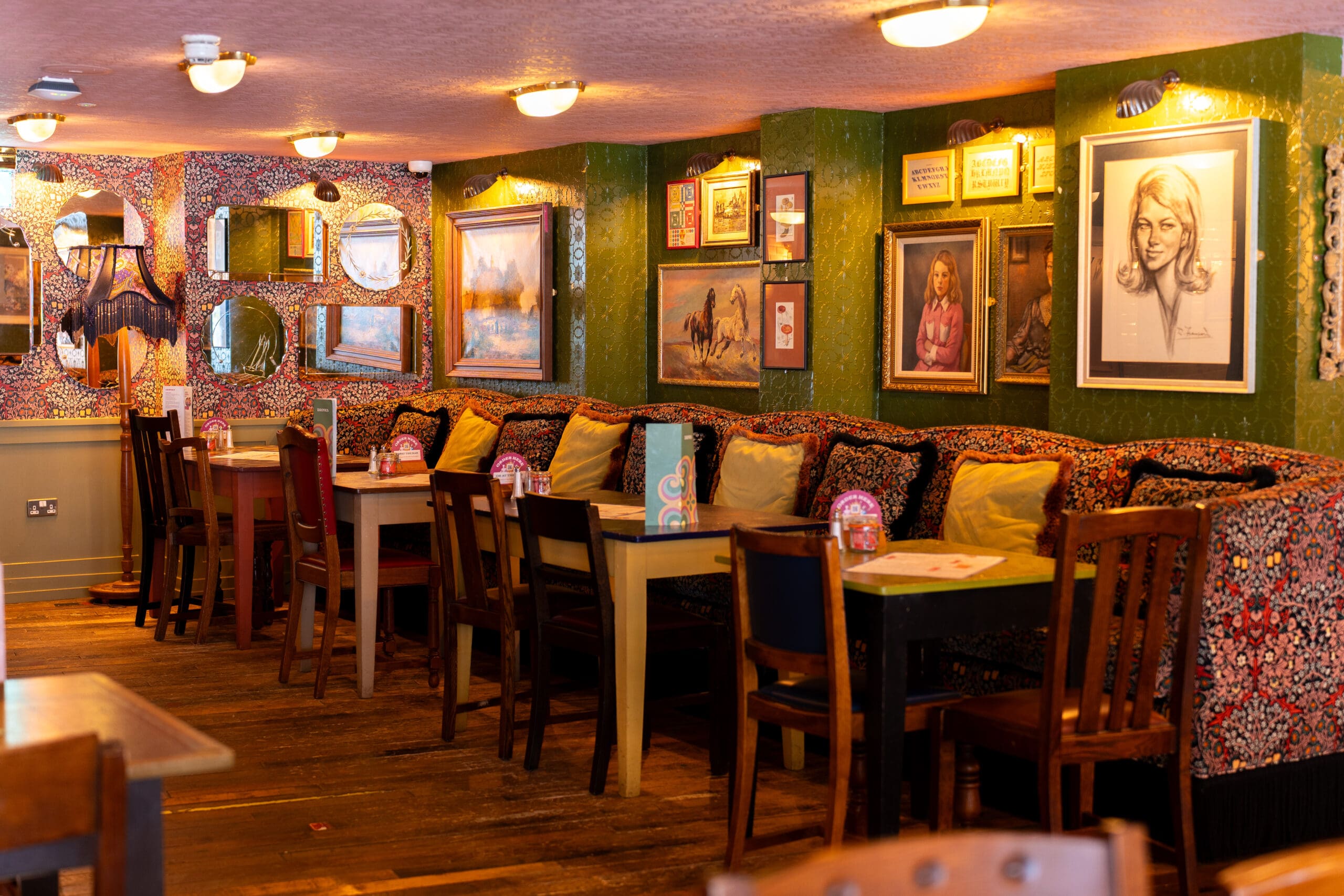 Cozy Bianco café interior with patterned benches, colorful cushions, wooden tables and chairs, mirrors, and framed artwork on green and floral walls. Soft lighting creates a warm, inviting atmosphere.