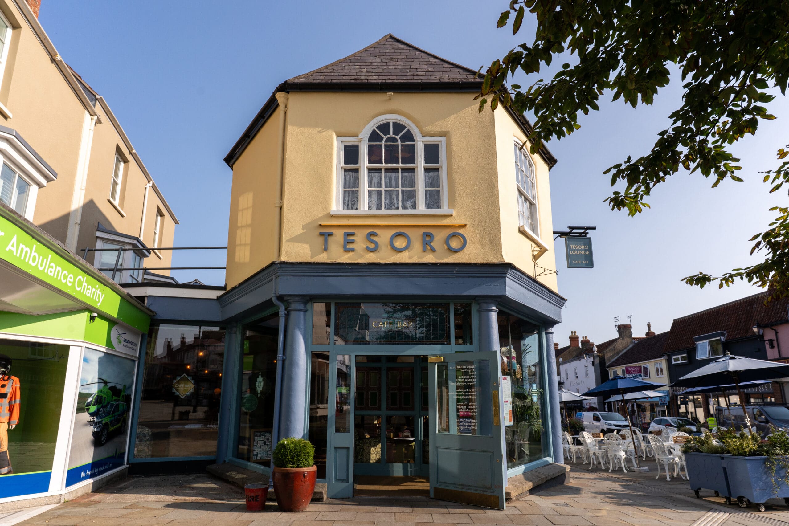 A yellow, two-story corner building with a sign reading "TESORO" above large Bianco-framed windows and a doorway. To the left is an ambulance charity shop, and outdoor seating is visible to the right.
