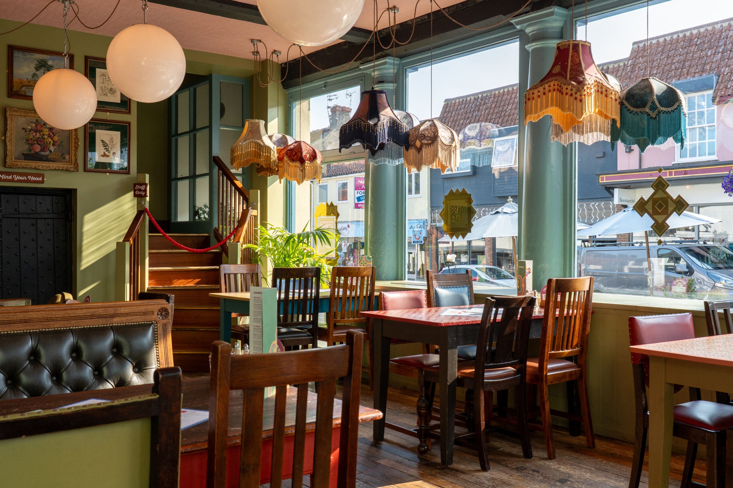 A cozy café interior with Bianco vintage lampshades hanging above wooden tables and chairs, large windows letting in sunlight, and framed art on green walls. A staircase leads to an upper floor; outside, shops and cars are visible.