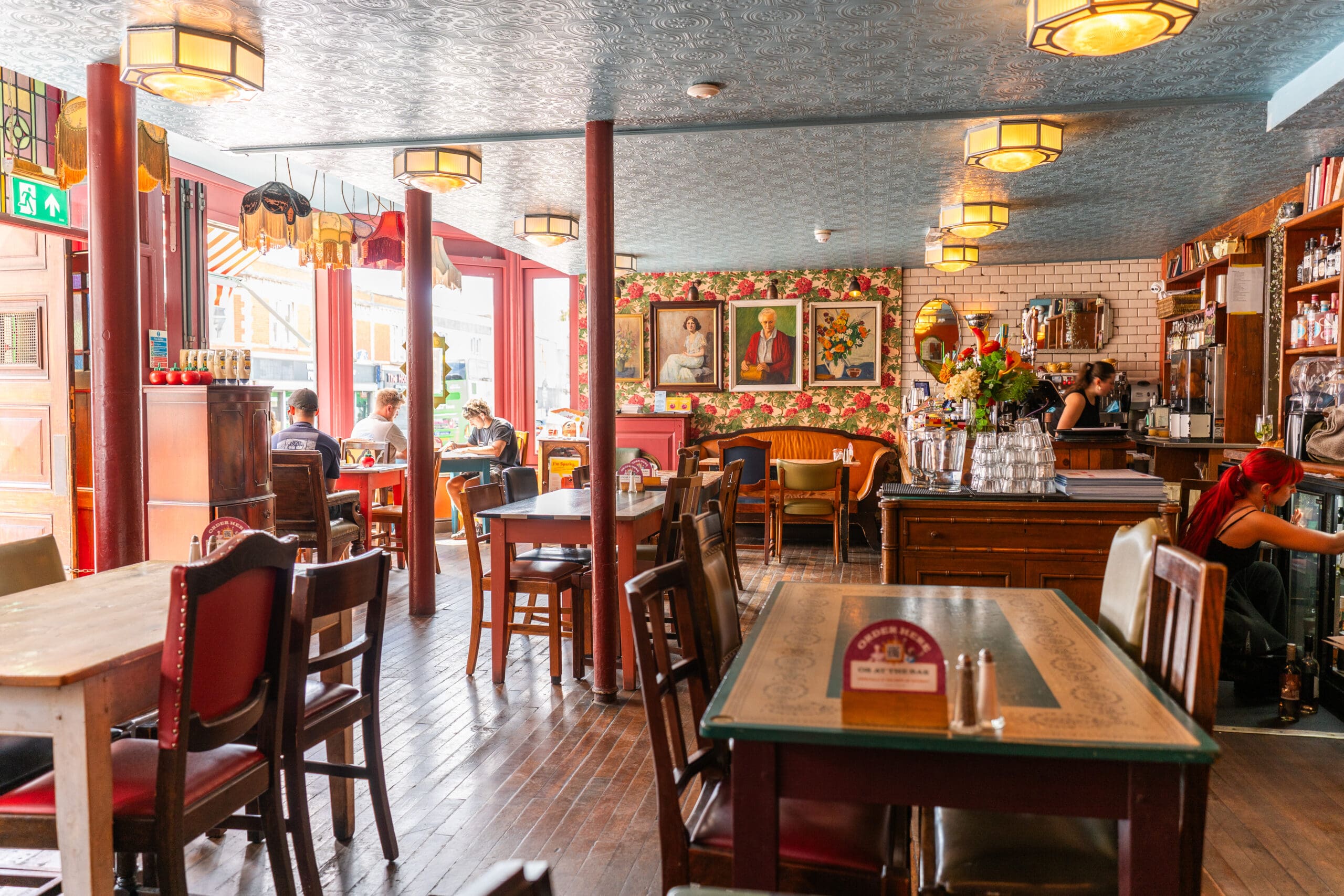 A cozy café interior with eclectic furniture, floral wallpaper, and framed portraits on the wall. Several people sip Tinto at tables and the counter, while large windows let in natural light from outside.