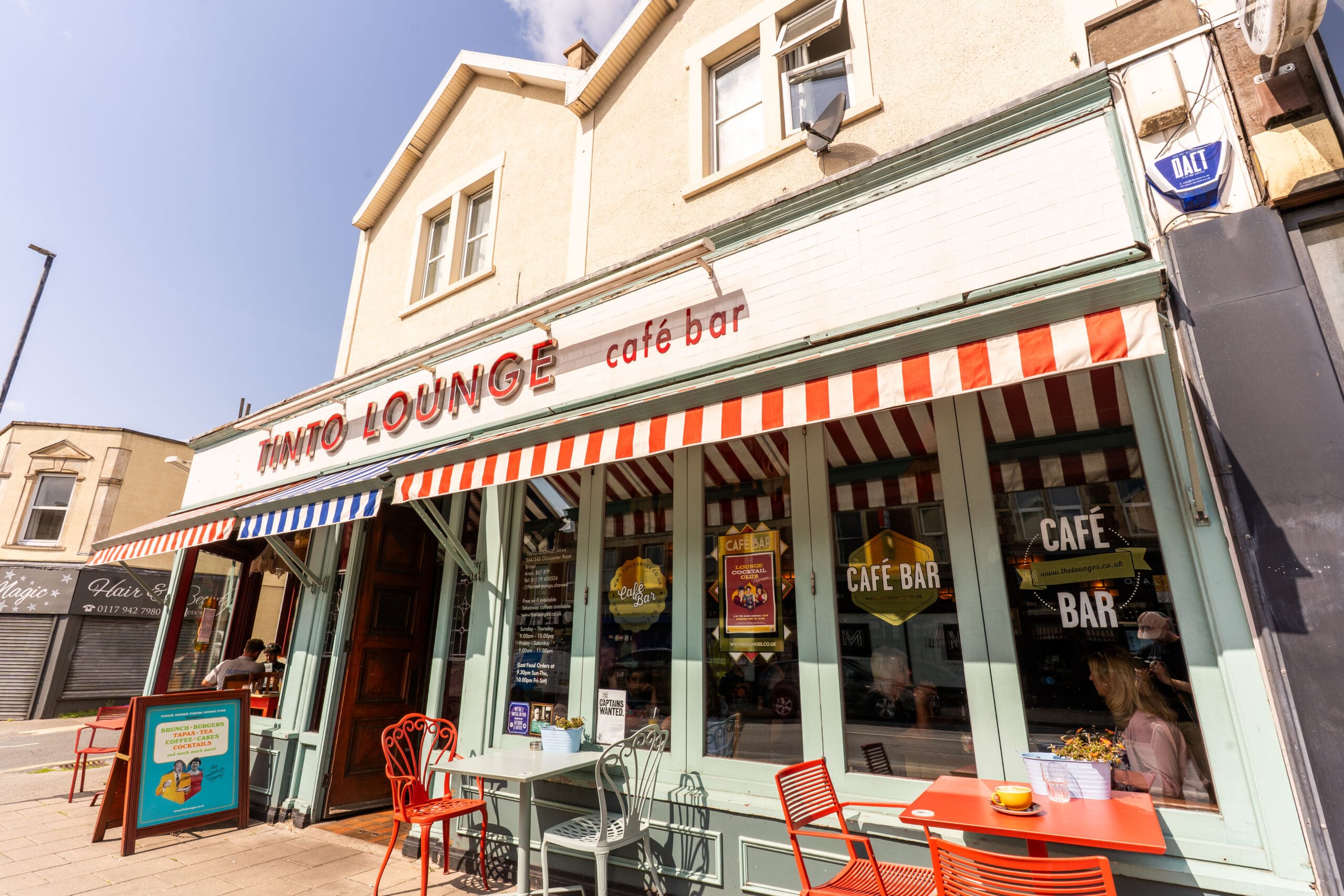 Tinto Lounge café bar, with its signature red and white striped awning, invites guests to relax at vibrant red tables and chairs under the sun. Large windows reflect the clear day as a person enjoys their coffee near the window at Tinto.