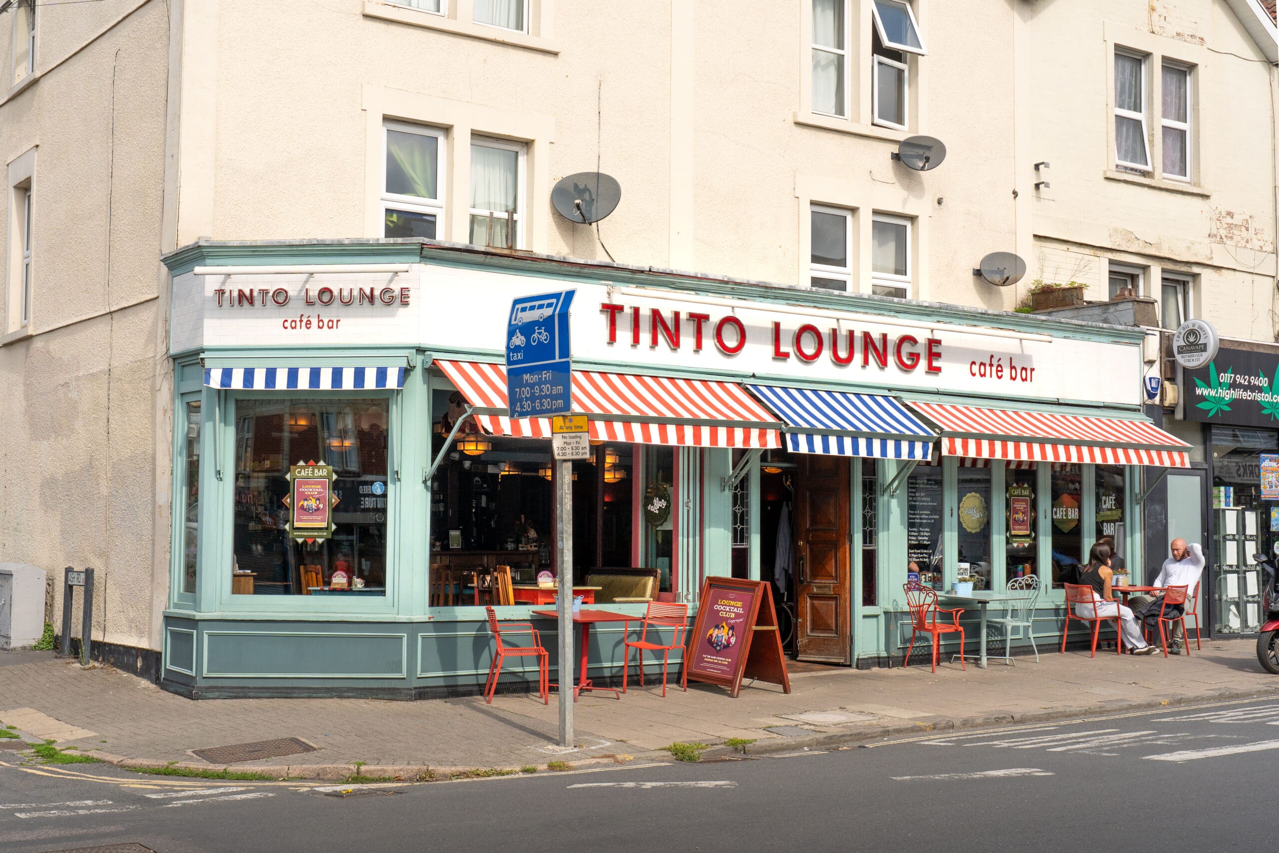Tinto Lounge café bar sits on a street corner with red, blue, and white striped awnings, outdoor tables and chairs, and people relaxing outside; a bicycle lane sign stands nearby, welcoming cyclists to Tinto’s vibrant setting.