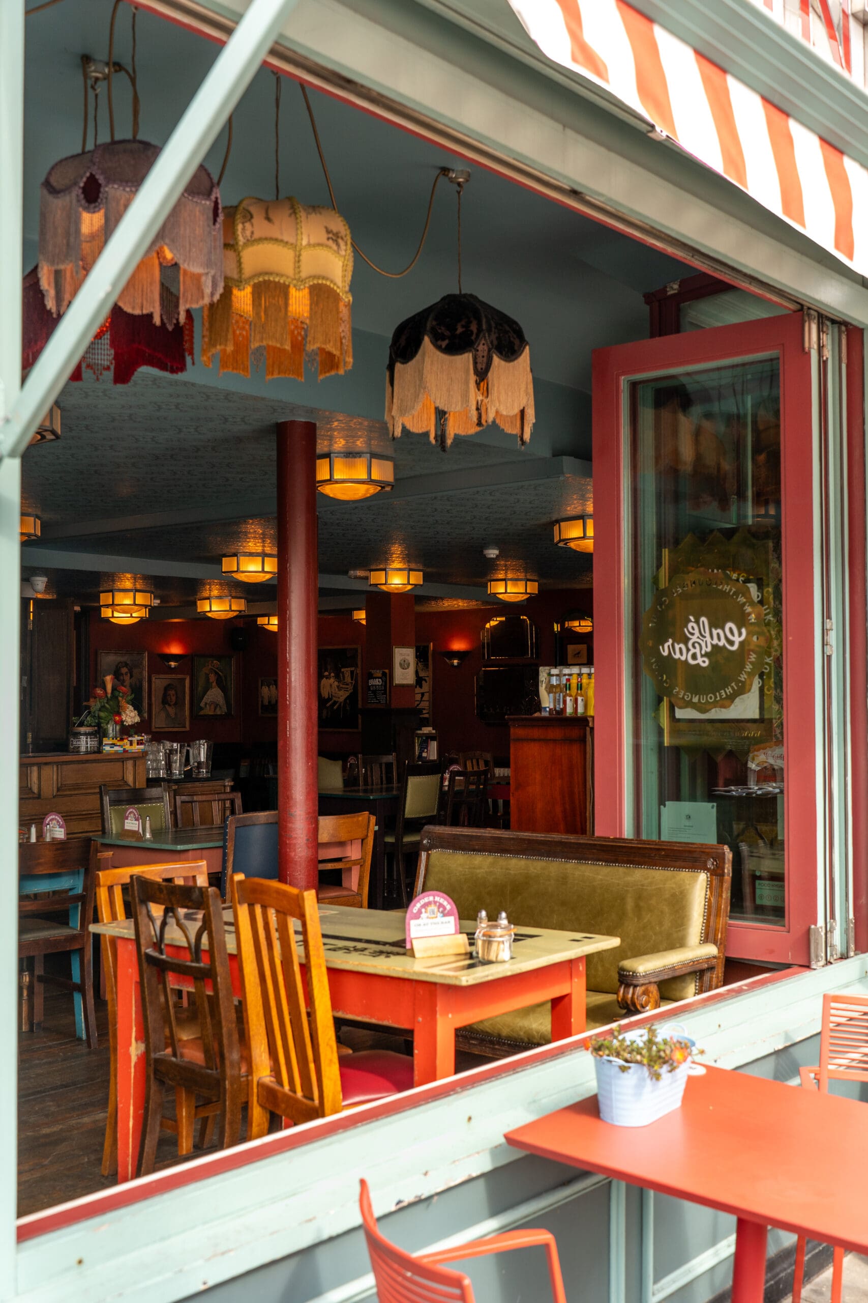 A cozy café interior with vintage décor, colorful chairs, wooden tables, and fringed lamps. The window is open, revealing a potted plant and a cup of Tinto on an outdoor table, adding to the welcoming atmosphere inside.