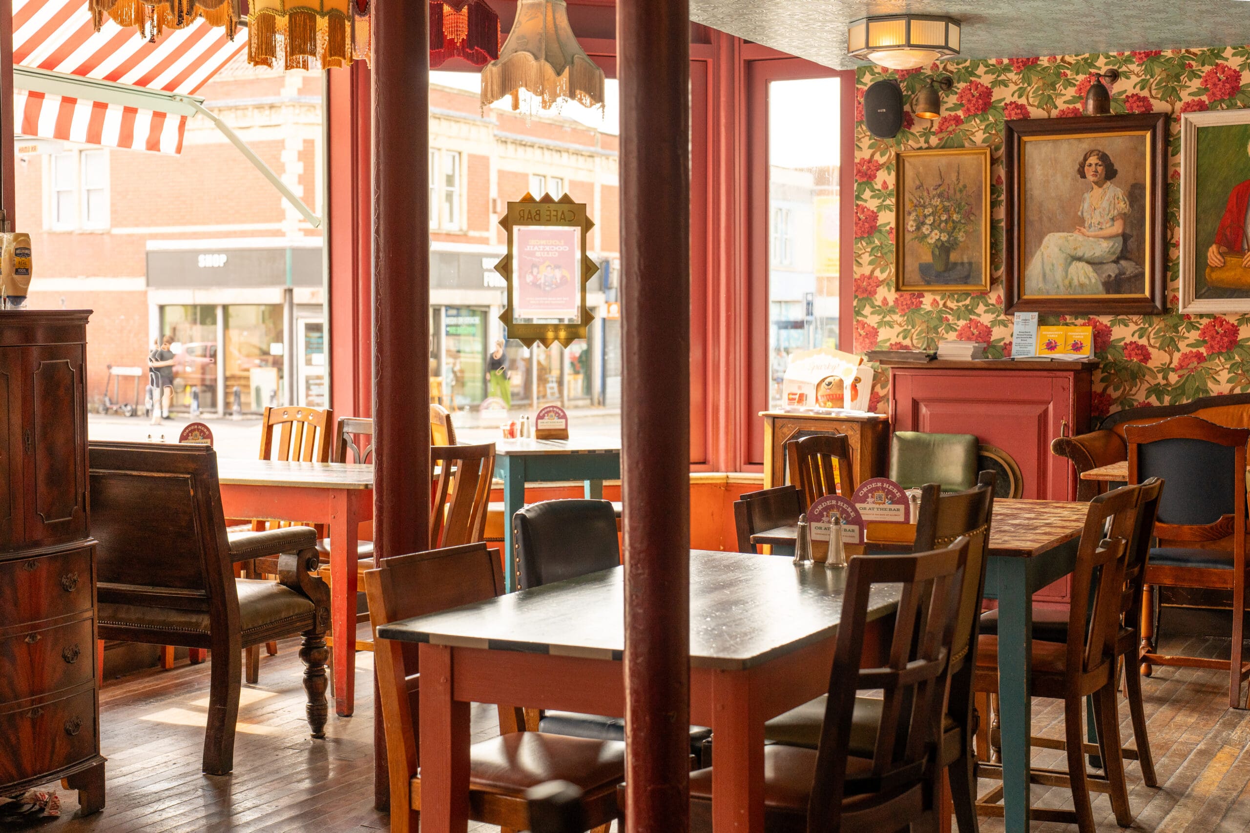 A cozy, vintage-style cafe with floral wallpaper, mismatched wooden chairs, hanging lamps, and framed paintings. Sunlight streams in through large windows as guests sip Tinto while admiring the charming street view outside.