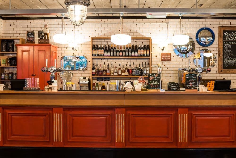 A bar with red cabinetry, wooden countertop, and shelves stocked with bottles. Hanging lights illuminate the white tiled wall, and a chalkboard menu is visible on the right side. Otto coffee equipment and taps are on the counter.