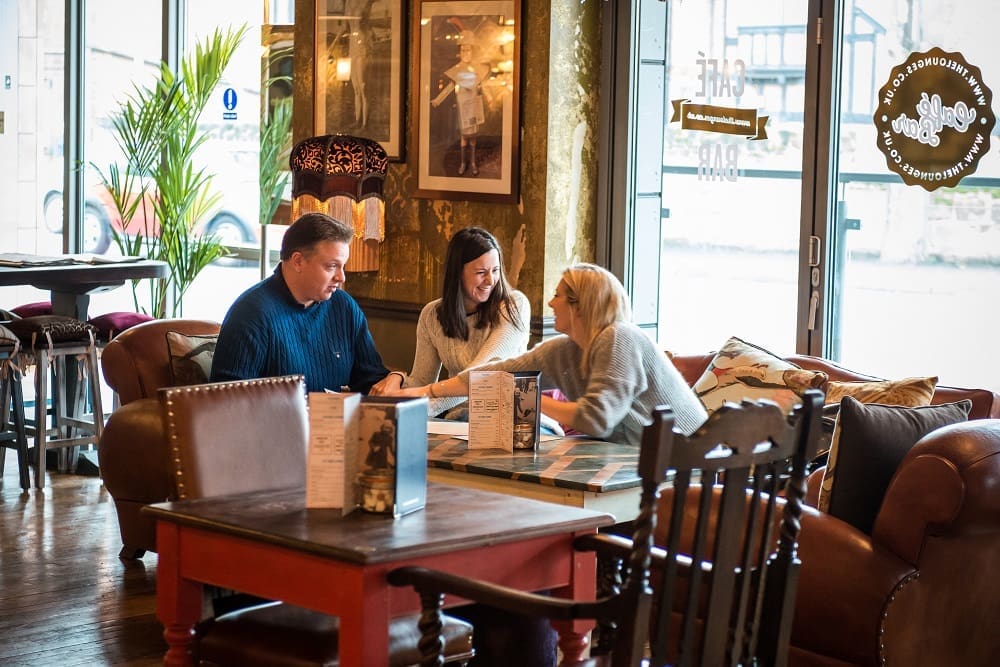 Three people sit together at a table in Otto, a cozy café, talking and looking at papers. The space features warm lighting, comfortable chairs, and large windows letting in natural light.