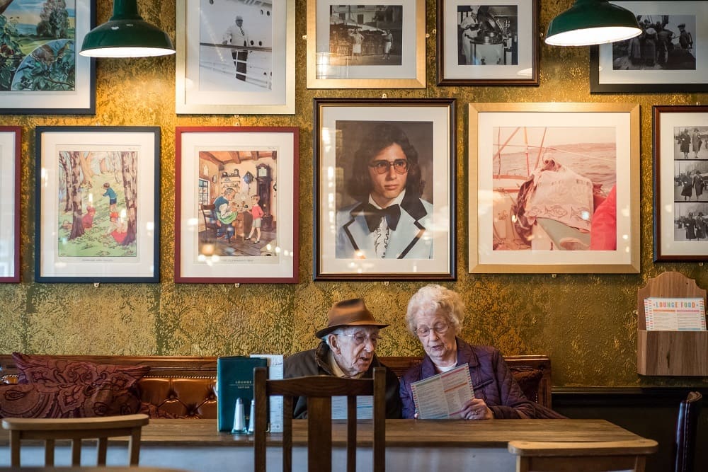 An elderly man and woman sit together at a table in Otto’s cozy cafe, reading a menu. Behind them, the wall is decorated with numerous framed photographs and artworks. Warm lighting creates a welcoming atmosphere.