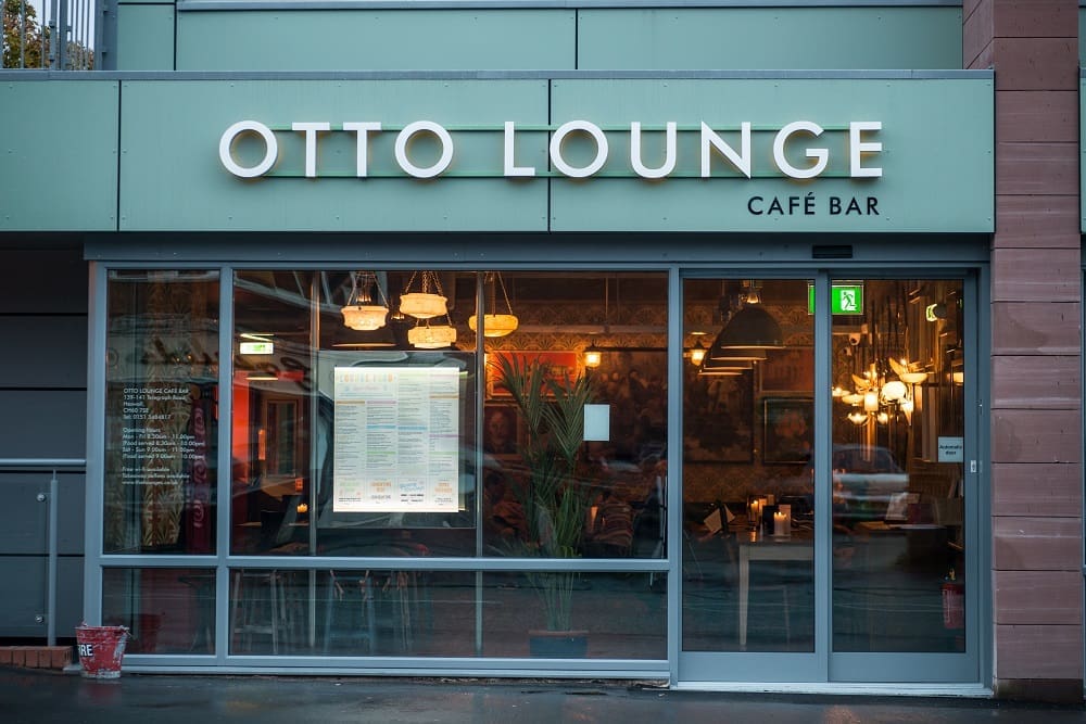 Exterior view of Otto Lounge Café Bar with large glass windows revealing warm indoor lighting, hanging lamps, a visible menu, and stylish interior decor. The street outside OTTO shines with reflections from the rain.