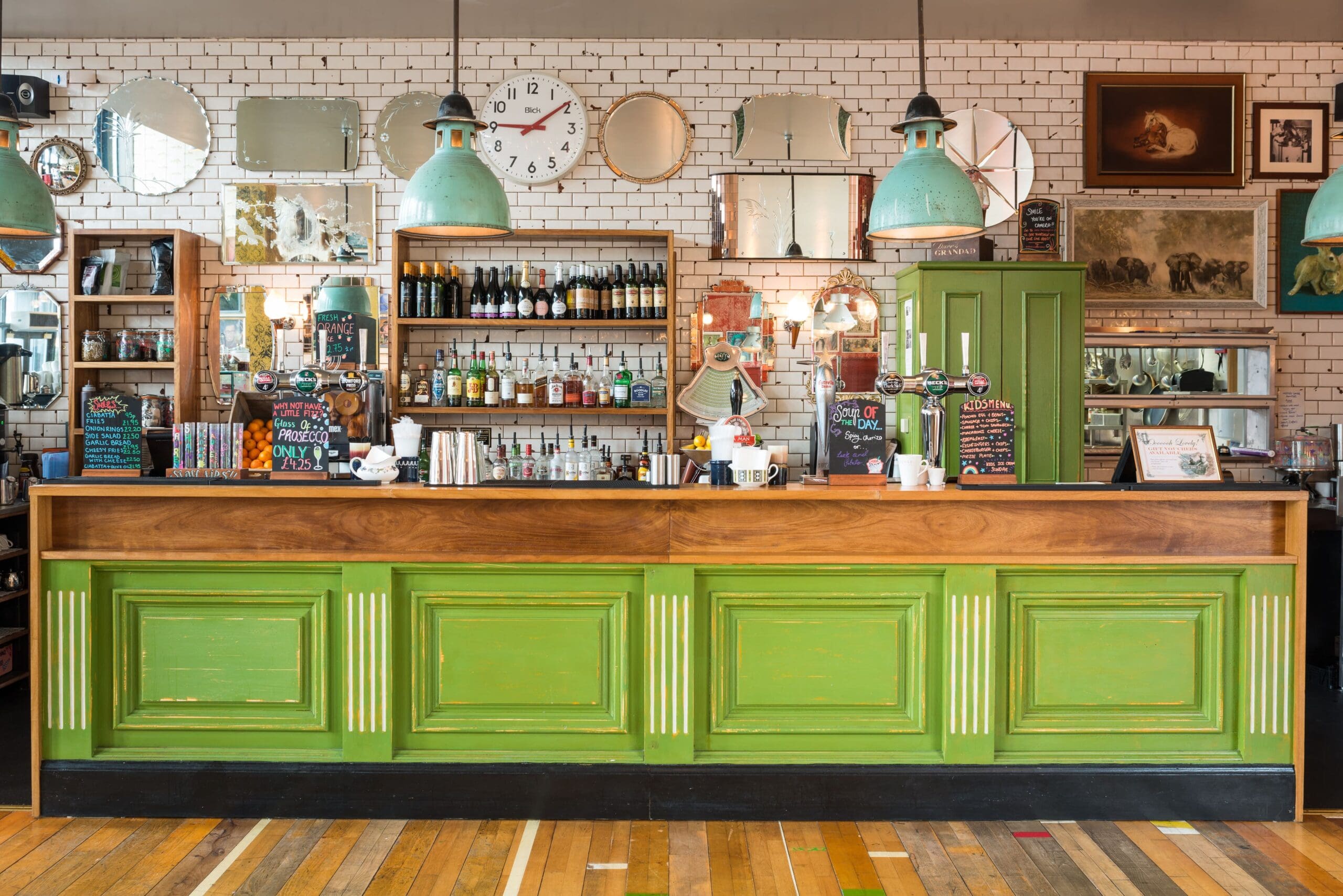 A vintage Pico pub bar with a bright green wooden counter, shelves of bottles, mismatched mirrors, clocks, and teal industrial pendant lights hanging from the ceiling. The decor features exposed brick and eclectic wall art.