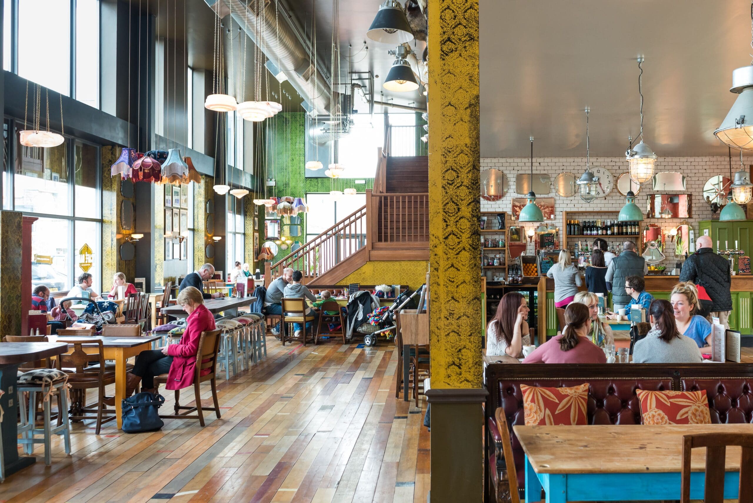 A spacious, brightly lit café with wooden floors, large windows, eclectic decor, and customers sitting at tables—chatting, eating, working on laptops—enjoying the vibrant Pico vibe. A staircase is visible in the background near the counter.