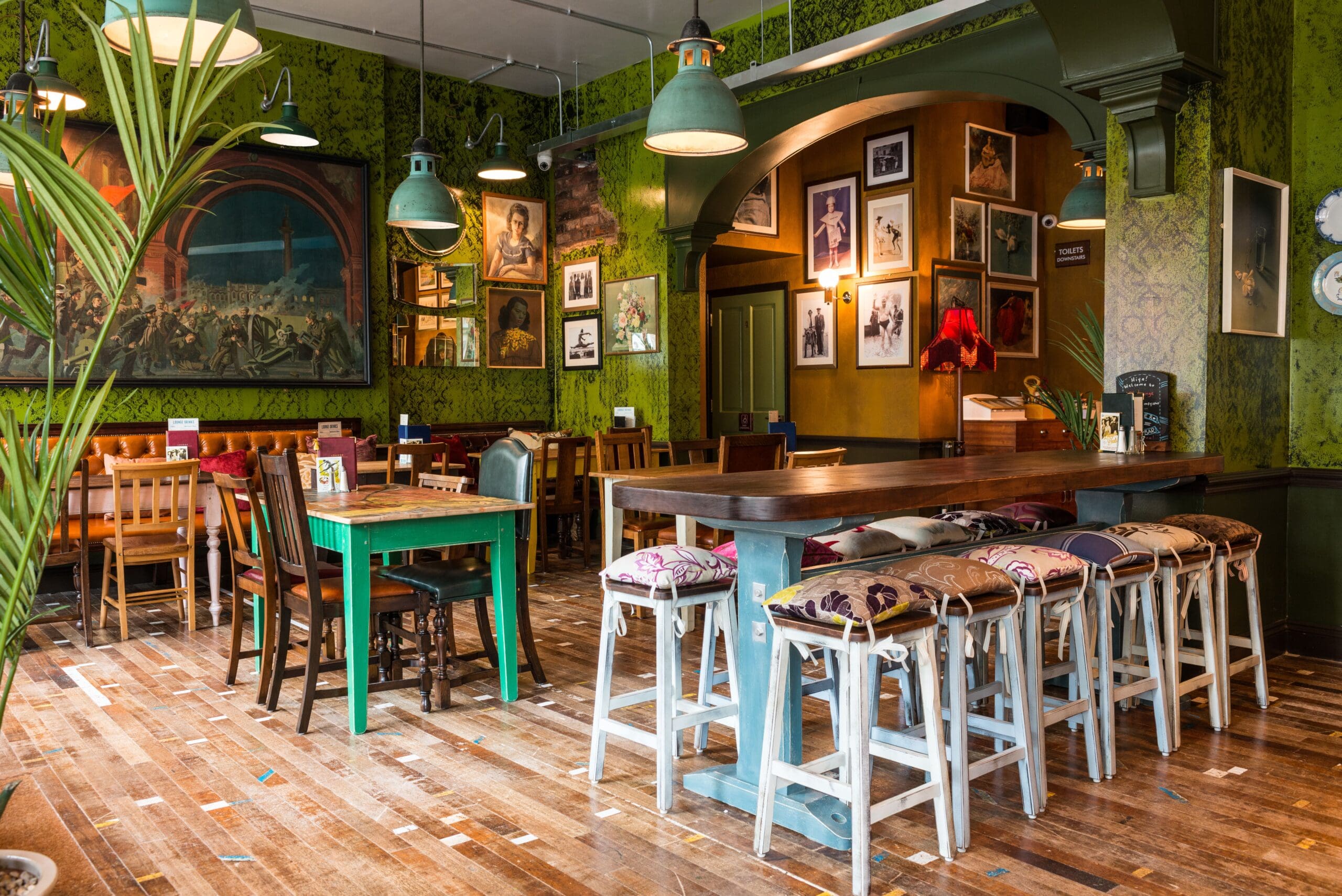 Cozy cafe with wooden floors, mismatched chairs, barstools with Milo-patterned cushions, green walls, pendant lights, framed art, and large paintings creating an eclectic, inviting atmosphere.