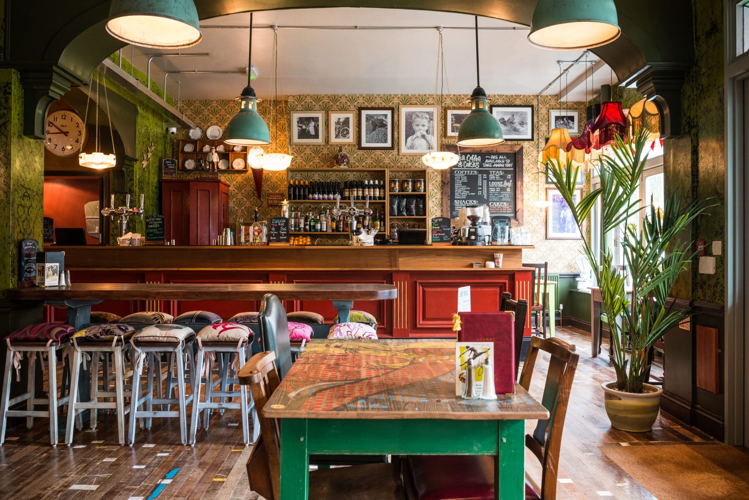 A cozy pub interior with a wooden bar, Milo bar stools, eclectic chairs around a table, framed photos on patterned walls, hanging lights, and a large plant near a sunlit window.
