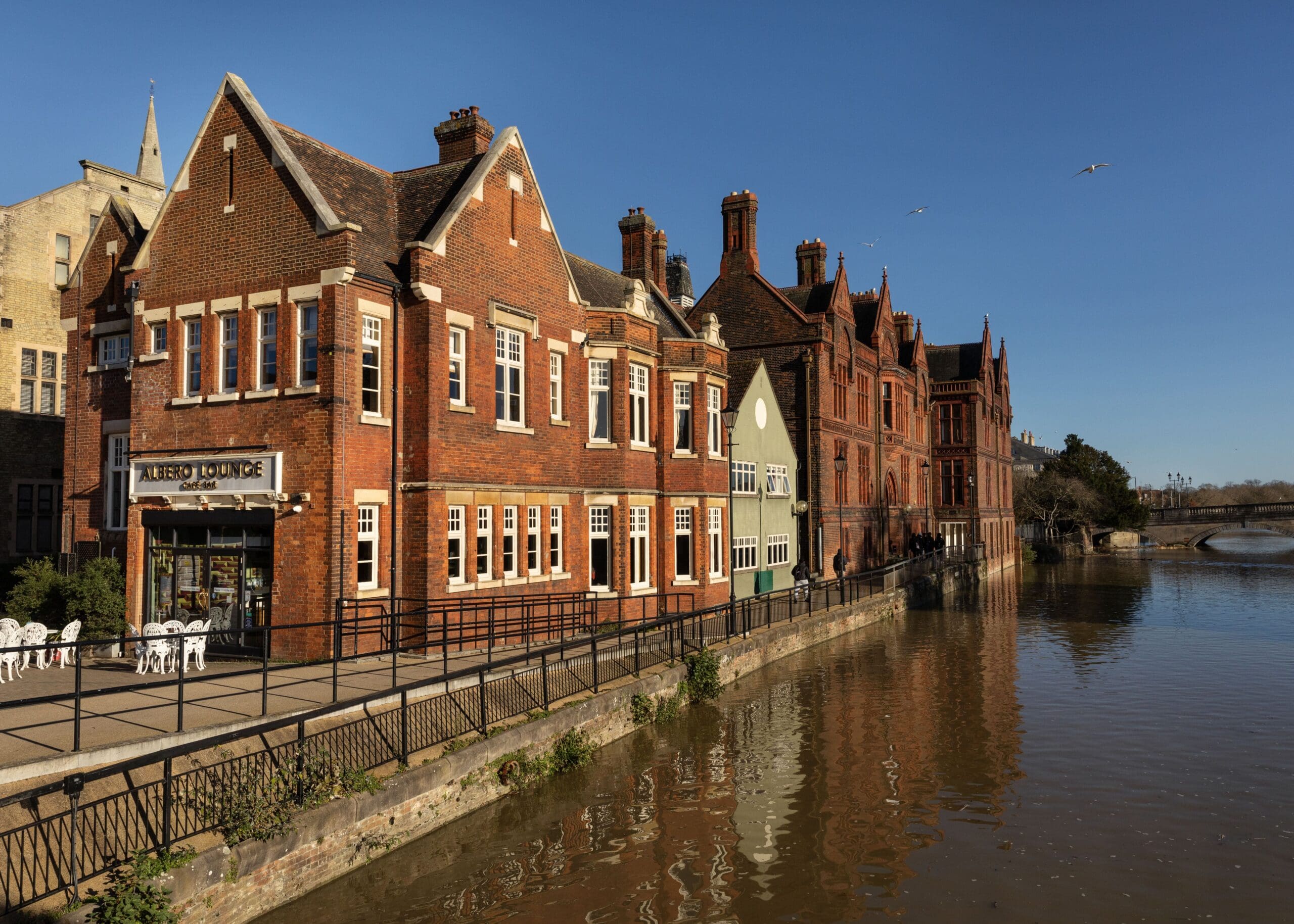 Red-brick buildings line the edge of a calm river under a clear blue sky, with Albero’s outdoor seating area and railings along the waterfront. A distant bridge and birds complete the picturesque background.