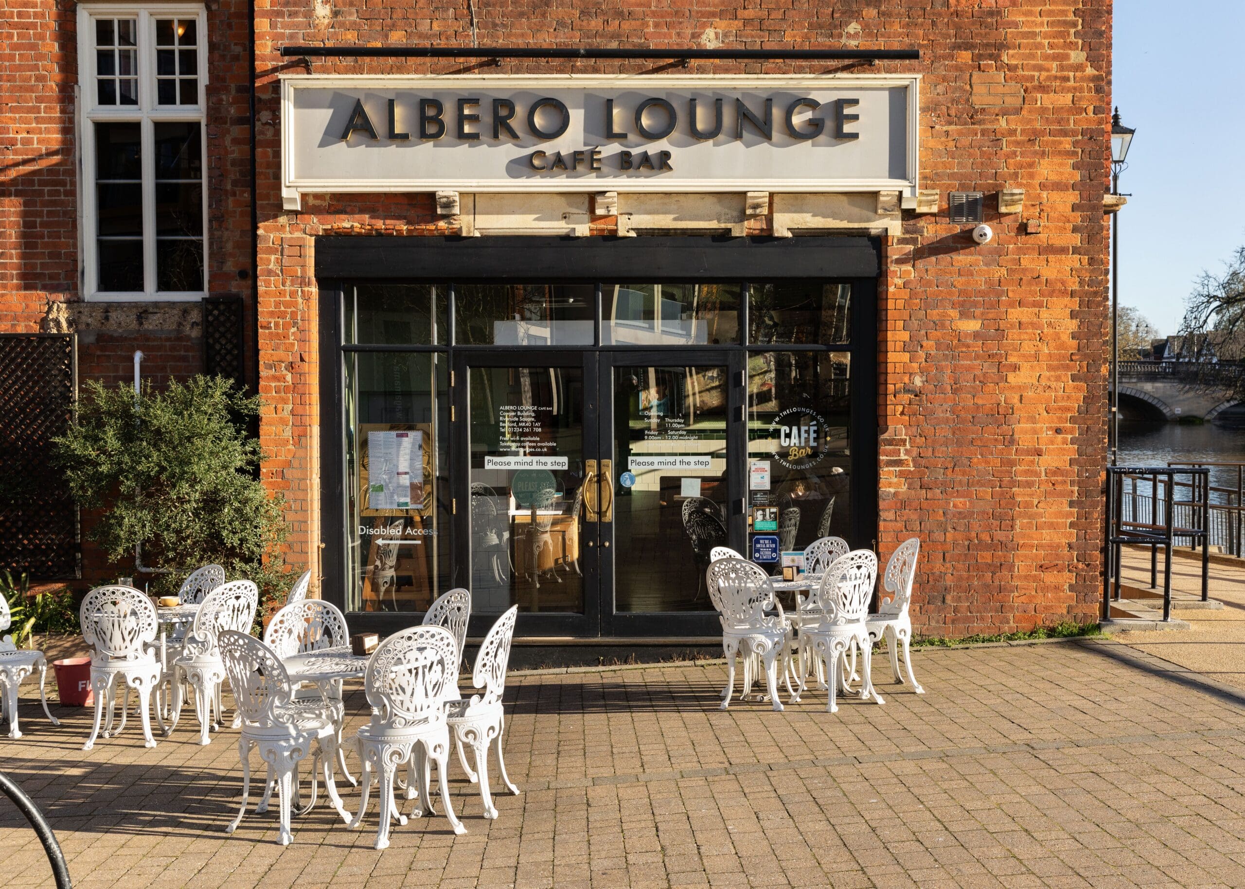Outdoor seating with ornate white chairs and tables is arranged in front of Albero Lounge Café Bar, a red-brick building with large windows and a sign above the entrance. The riverside setting adds to Albero’s inviting charm.