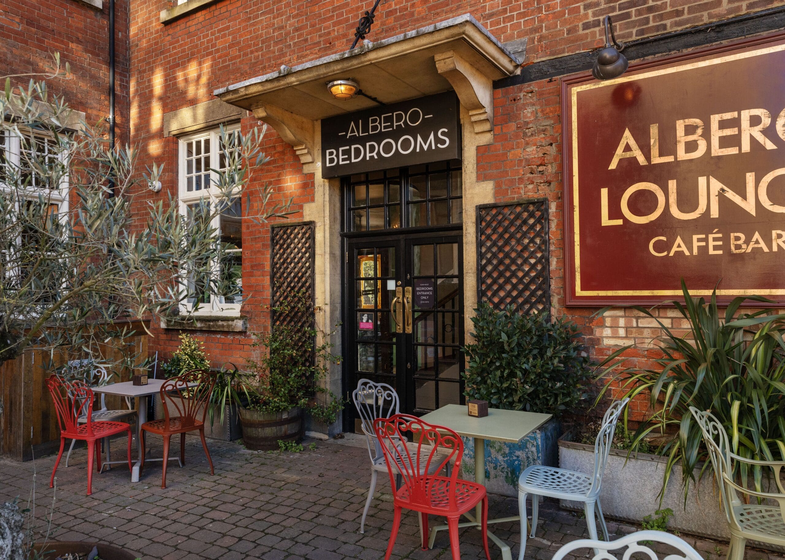 Outdoor seating area with colorful chairs and tables in front of a brick building. Signs read "Albero Bedrooms" above a door and "Albero Lounge Café Bar" on the wall, welcoming guests to experience Albero’s vibrant charm. Potted plants decorate the entrance.