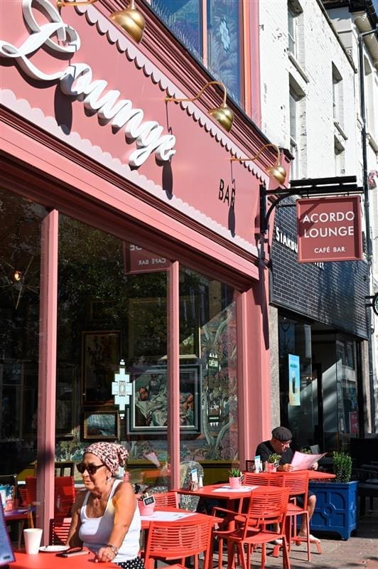 People sit at red tables outside Acordo on a sunny day. The café’s pink exterior, large windows, and sign above the entrance invite guests to relax while enjoying the vibrant street with other storefronts nearby.