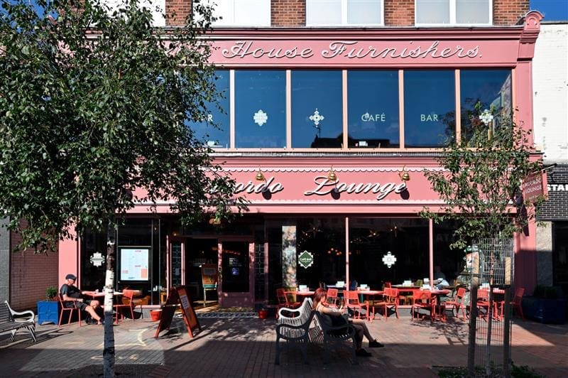 A pink, two-story café and bar called "Lounge" with large windows, red outdoor chairs and tables, where people enjoy an Acordo coffee outside under trees on a sunny day.