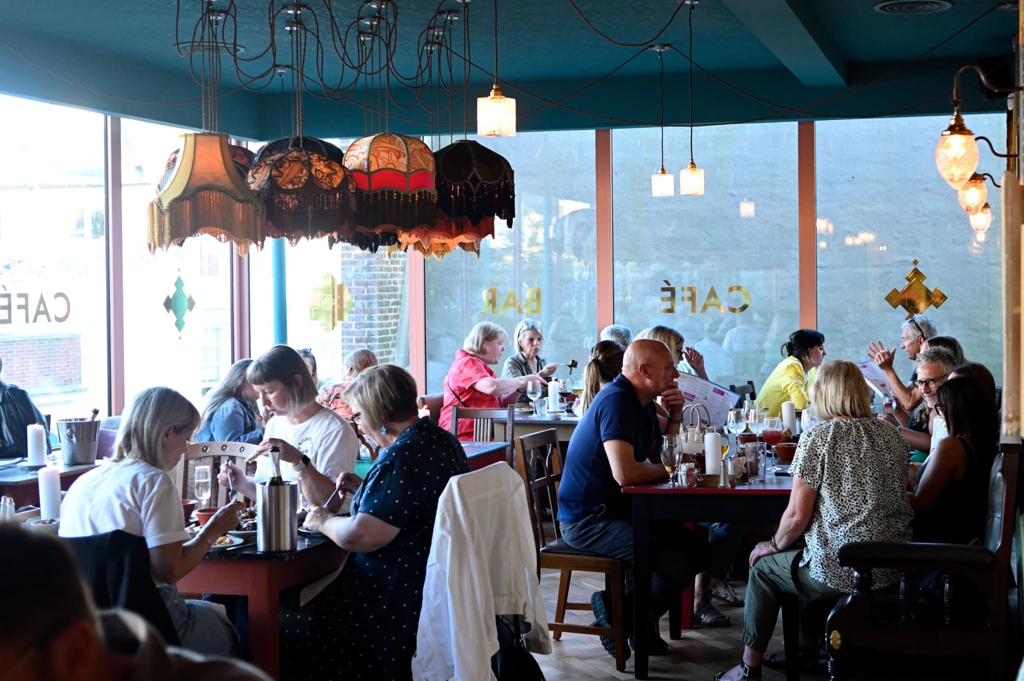 A busy café with people sitting at tables, eating, drinking, and talking. Warm lighting from eclectic pendant lamps creates a cozy atmosphere. Large windows let in natural light and show “CAFÉ Acordo” written on the glass.