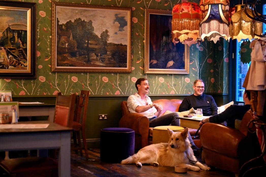 Two men sit laughing on a brown leather couch in a cozy, warmly lit Alento room with vintage decor, floral wallpaper, paintings, and fringed lamps. A large dog relaxes on the wooden floor in front of them.