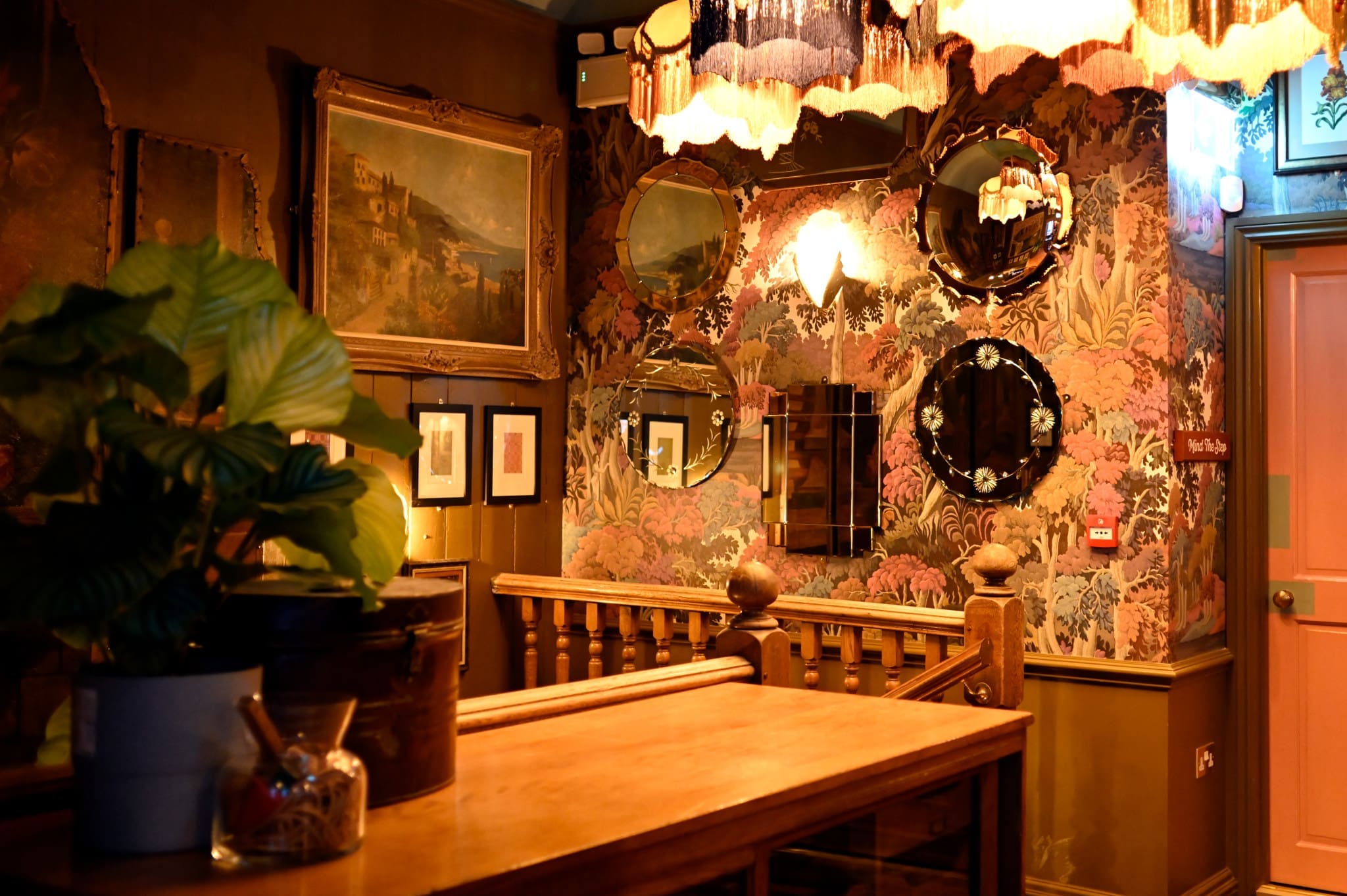 A warmly lit, vintage-style interior with patterned wallpaper in rich Bordo hues, ornate mirrors, framed artwork, hanging lamps, and potted plants on a wooden counter near a staircase and a pink door.