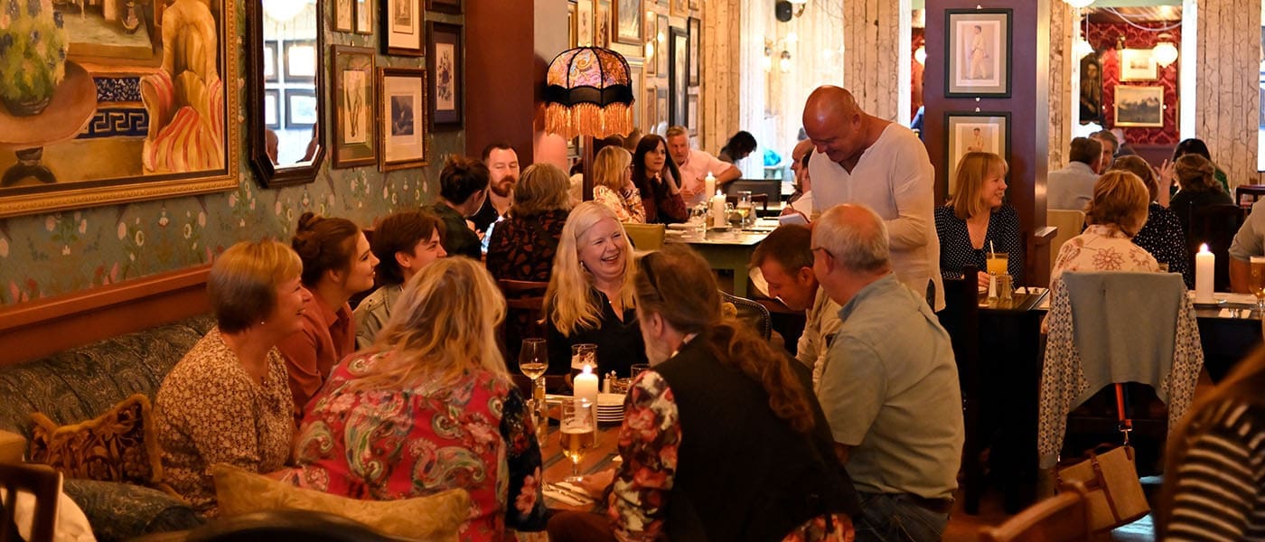 A group of people sit around a table in a cozy, dimly lit restaurant, chatting and laughing. The walls feature eclectic artwork and vintage lamps by Alturo, creating a warm and inviting atmosphere.
