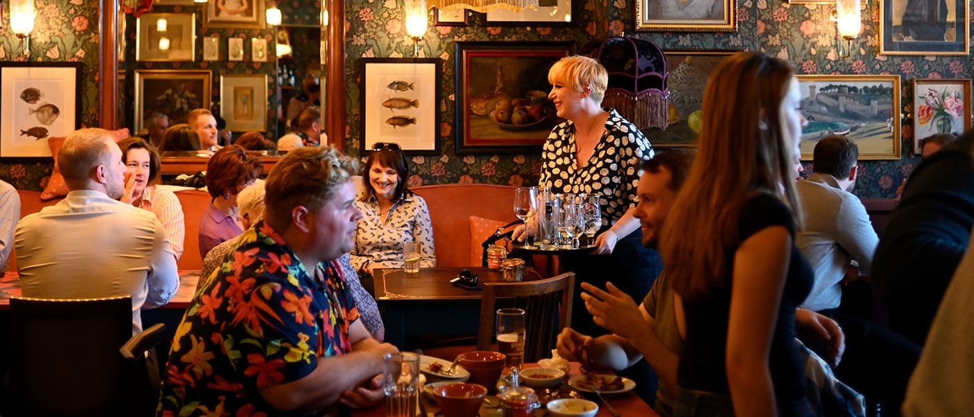 A lively restaurant, Alturo, with people eating, drinking, and talking at tables. A server stands with a tray of glasses. The walls feature various framed artworks and patterned wallpaper, creating a cozy, vibrant atmosphere.