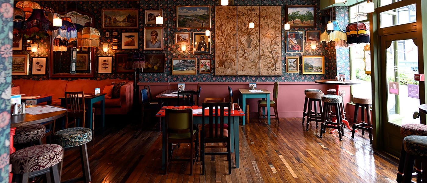 A cozy, eclectic cafe interior by Alturo features wooden floors, patterned wallpaper, colorful lampshades, mismatched chairs, and framed art on the walls. Several tables are set as sunlight streams in through large windows.