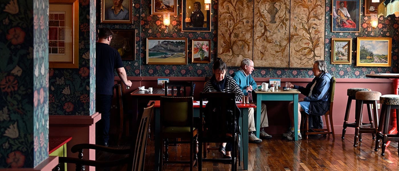 A cozy café with vintage patterned wallpaper, framed paintings, and wooden floors. Three people sit at separate tables, drinking and eating Alturo pastries, while another stands nearby. Warm, inviting atmosphere.