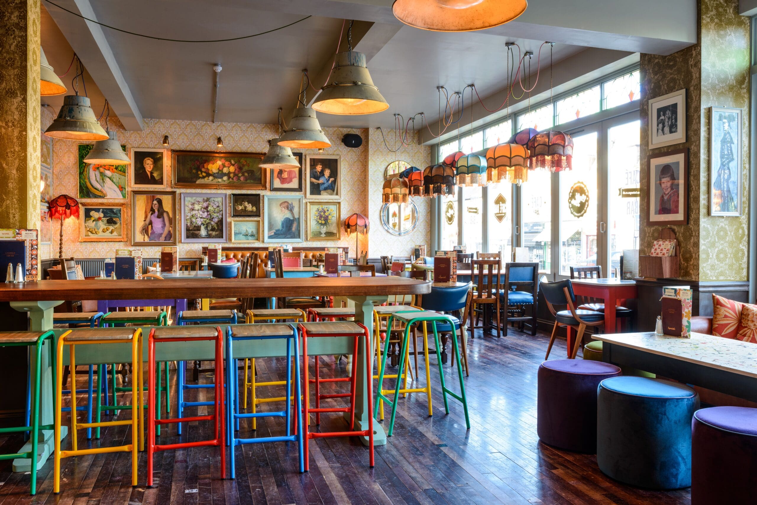 A colorful, eclectic cafe near the Metro, featuring rainbow-colored bar stools, vintage lamps, wooden tables, and a gallery wall of assorted paintings. Large windows fill the space with natural light for a cozy, inviting atmosphere.