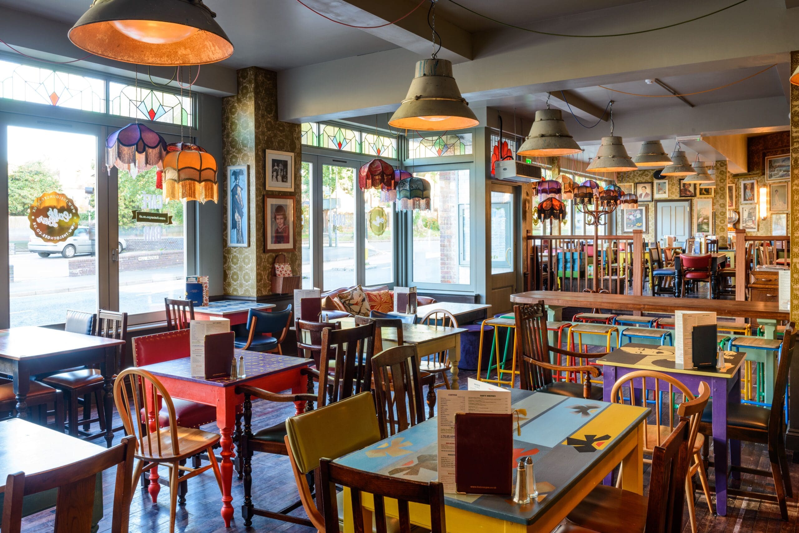 Colorful, eclectic café interior with mismatched chairs and tables, large windows letting in natural light, metro-inspired vintage lamps hanging from the ceiling, and quirky wall art and decorations throughout the space.