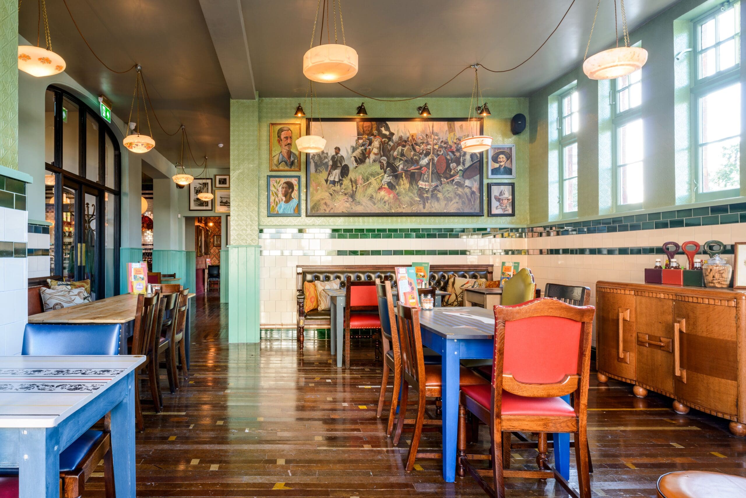A brightly lit restaurant with colorful chairs, wooden tables, tiled walls, hanging lamps, framed artwork, and large windows letting in natural light. The inviting atmosphere pairs perfectly with Albero’s eclectic charm.