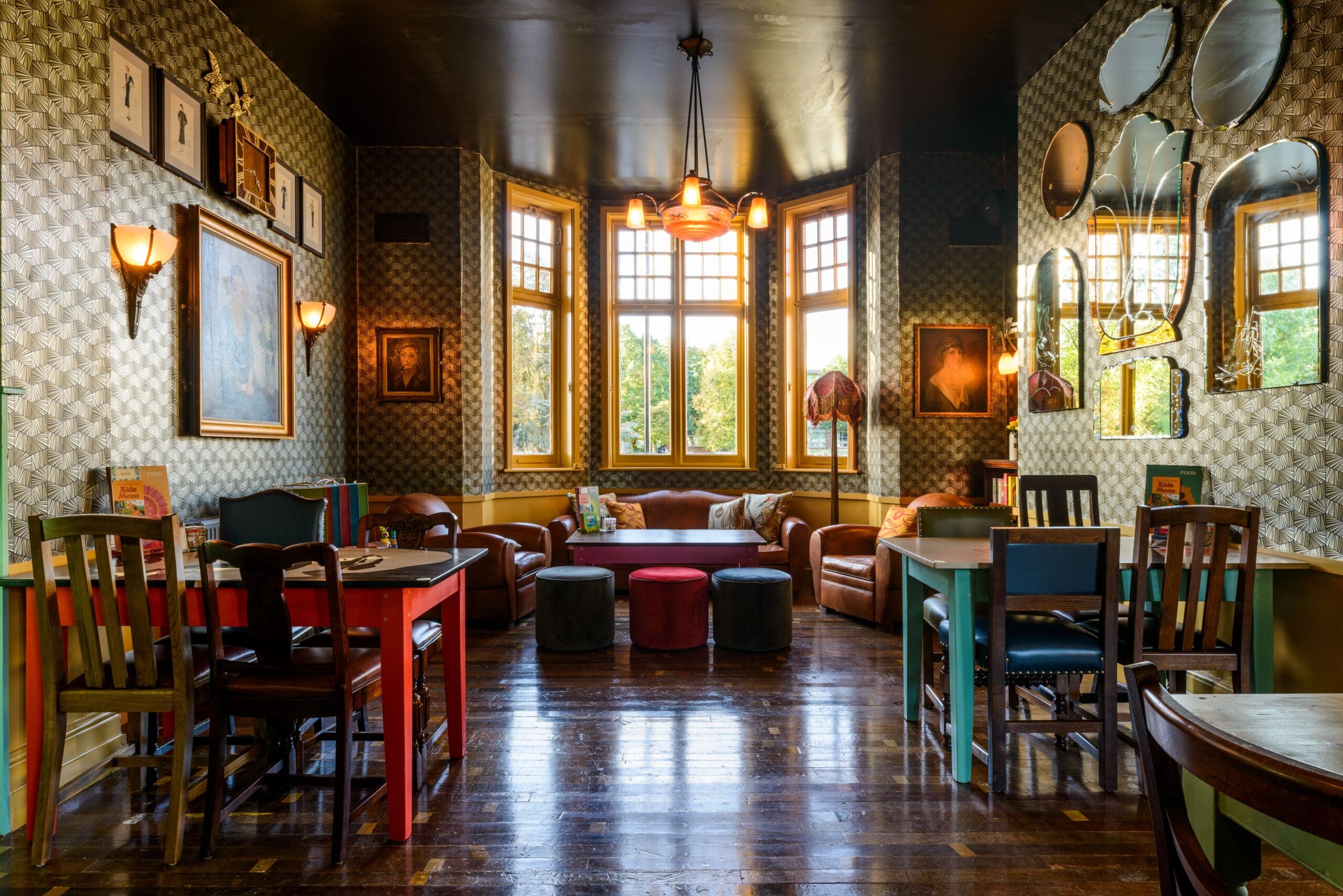 Cozy, stylish Albero café interior with wooden tables and chairs, leather sofas by large bay windows, patterned wallpaper, framed art, round mirrors, and warm lighting reflecting on polished wooden floors.