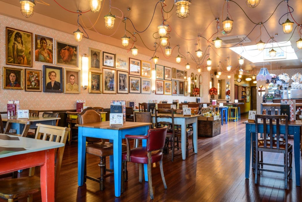 A brightly lit, eclectic restaurant interior features colorful mismatched tables and chairs, wooden floors, striking Arco pendant lights, and walls adorned with framed vintage portraits and photos.