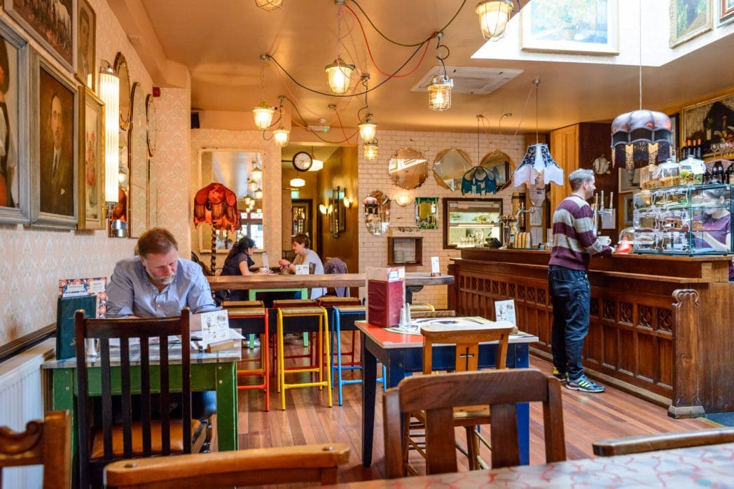 A cozy café with colorful mismatched furniture, quirky lamps—including an elegant Arco lamp—and paintings on the walls. People sit at tables reading or talking, while a person stands at the wooden counter near a pastry display. Warm, inviting atmosphere.