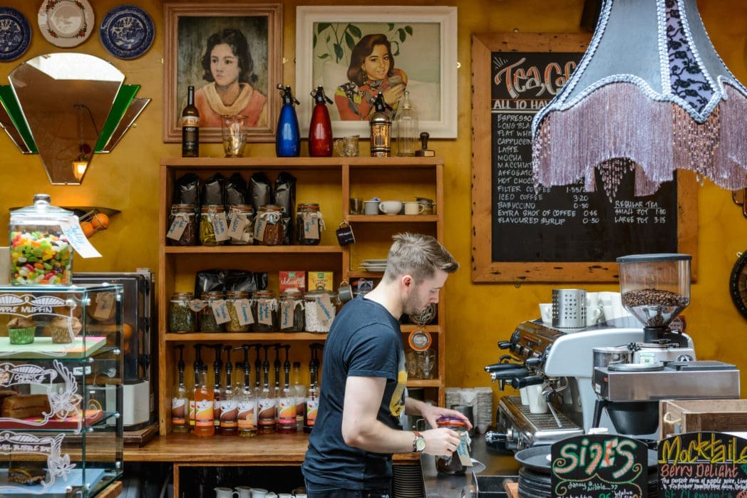 A barista prepares drinks behind the counter of a cozy café, surrounded by Arco coffee equipment, jars, bottles, and tea canisters. Colorful decor and framed portraits hang on the yellow wall above.