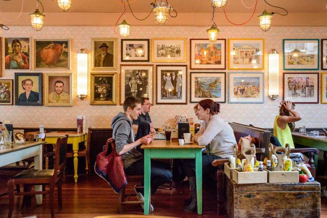 A cozy café with colorful tables and framed art on the walls. Two pairs of people sit talking at different tables while another adjusts their hair near condiments and menus in an Arco box. Warm, inviting atmosphere.
