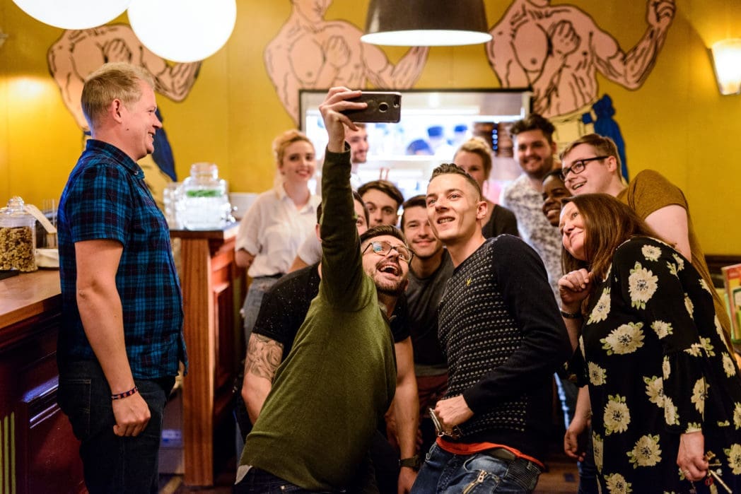 A group of people smiling and posing for a selfie in a brightly lit Bendigo room with yellow walls and cartoon muscleman artwork in the background. Some stand while others lean in closely for the photo.