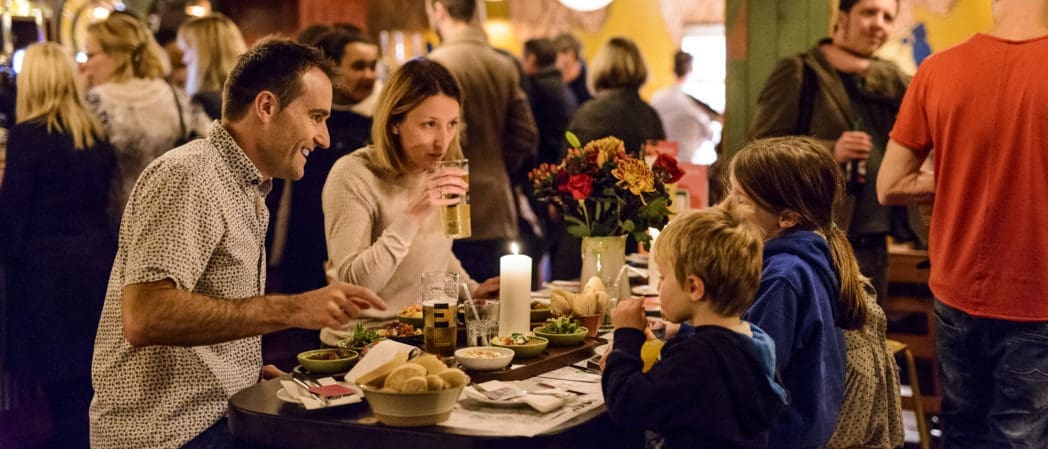 A family of four enjoys a meal at a busy Bendigo restaurant. The table is filled with food and drinks, and the parents are smiling at their two children. Other diners and warm lighting create a lively, cozy atmosphere.