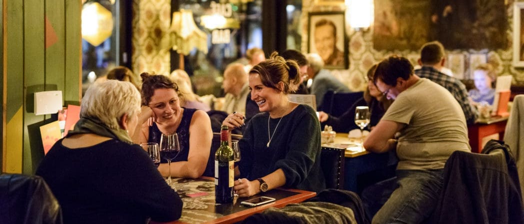 A group of people sit at tables in a cozy, dimly lit Bendigo restaurant, enjoying drinks, wine, and conversation. Some people are laughing and smiling, creating a warm and lively atmosphere.