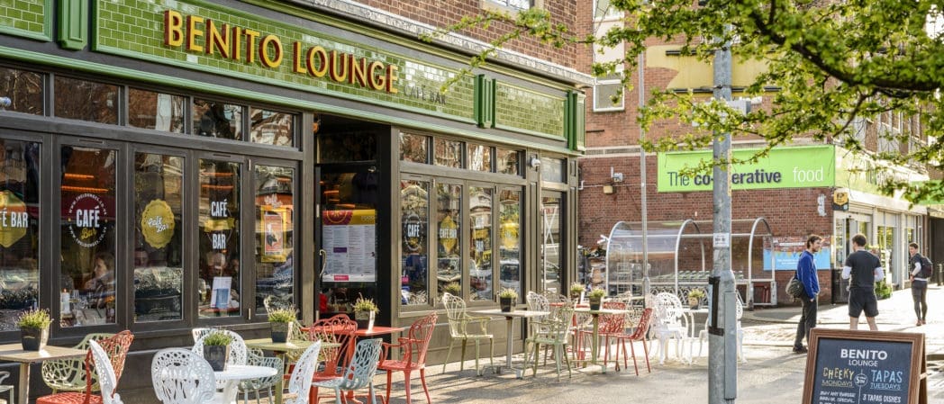 Outdoor seating with colorful chairs and tables in front of Benito Lounge café, located on a city street. People walk by Benito’s inviting terrace, while a Cooperative Food store is visible across the road. The scene is lively and sunny.