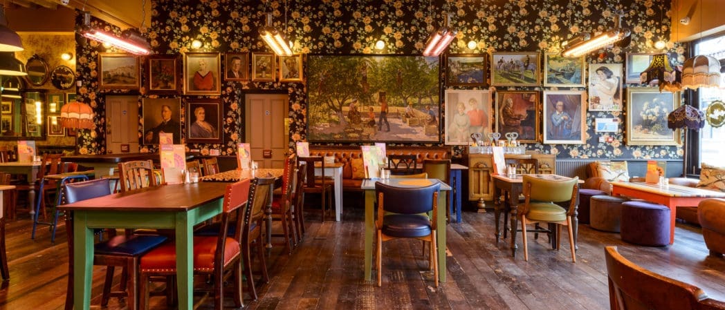 A cozy, eclectic café with colorful chairs, wooden tables, floral wallpaper, and walls adorned with various paintings and framed art—Benito’s warm lighting creates an inviting atmosphere.