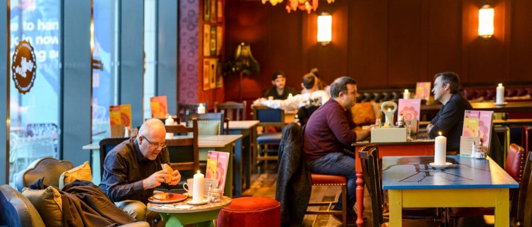Several people, some wearing a berretto, sit at tables in a cozy cafe with warm lighting. Some are eating or drinking, while others chat. Large windows on the left let in daylight, and menus and candles decorate the tables.