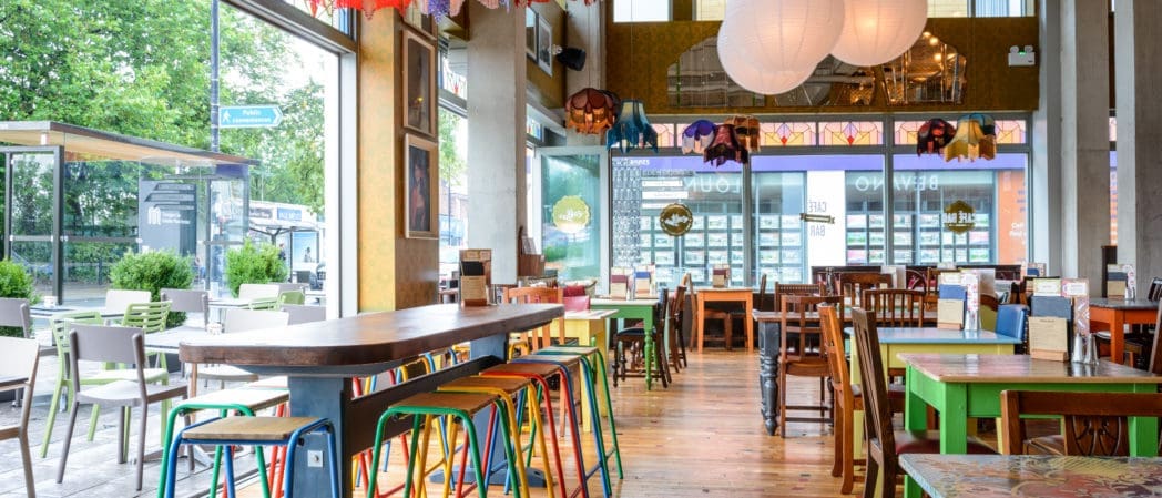 A colorful, modern café with vibrant chairs, wooden tables, and large Bevano paper lanterns hanging from the ceiling. Floor-to-ceiling windows let in natural light and reveal greenery outside. The space has an eclectic, welcoming atmosphere.