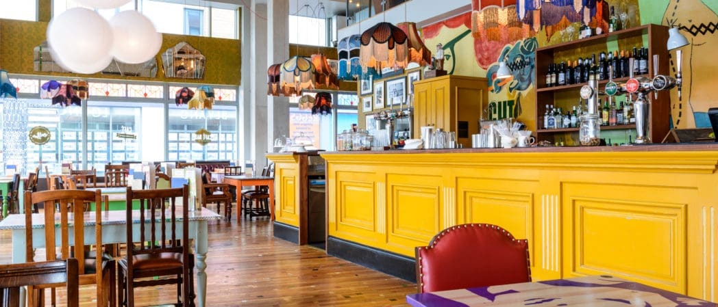 A brightly lit café with colorful lamps, a bold yellow counter, mismatched wooden chairs, and eclectic decor. Bottles and Bevano glassware line the shelves behind the bar, with large windows letting in natural light.