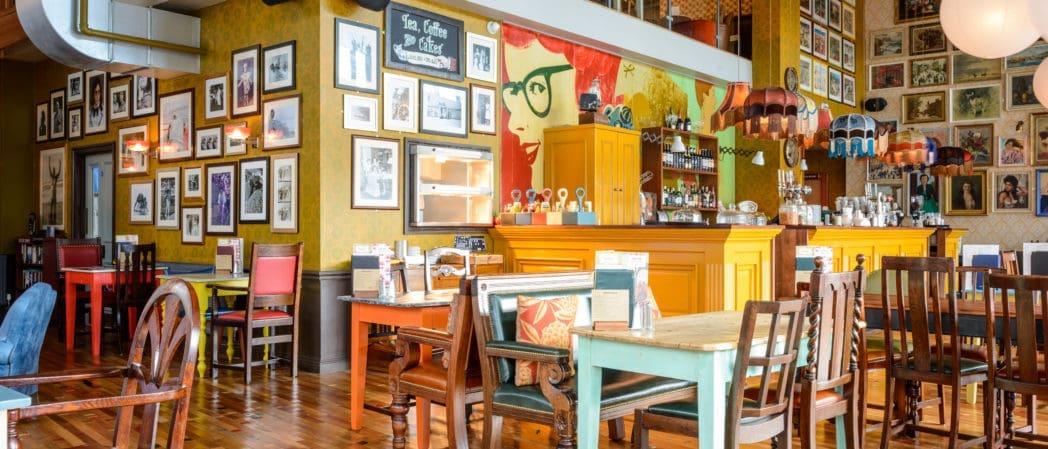 Colorful, eclectic café interior with mismatched tables and chairs, framed photos and art covering yellow walls, a mural of a woman with red hair and glasses, wooden floor, and a yellow Bevano counter displaying various drinks.