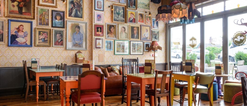 A cozy café, Bevano, with mismatched colorful chairs and tables, a brown leather sofa, vintage lamps, and a wall of framed portraits. Large windows flood the space with natural light, creating a warm, inviting atmosphere.