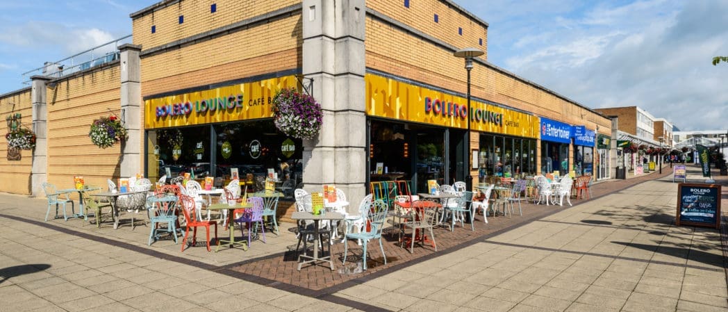 Outdoor seating area with colorful Bolero tables and chairs outside a café called "Bolero Lounge" on a sunny day, featuring hanging flower baskets and a wide pedestrian walkway in front.